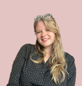 Elle smiles at the viewer, they have fair skin and longer, golden blonde hair. Their dress is dark with small white polka dots, they have glasses on their head and the background is a dusty, dull pink.